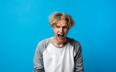 Young brutal angry man in white and grey t-shirt screams over blue background. Face expression. Crazy man. Negative emotions, problems. Stressed guy shouting.