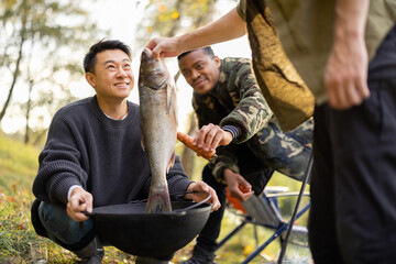 European man putting big fish in cauldron for cooking Ukha soup while his multiracial friends...