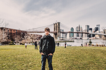 Young adult man standing in brooklyn on a meadow looking at camera, brooklyn bridge and skyline of...