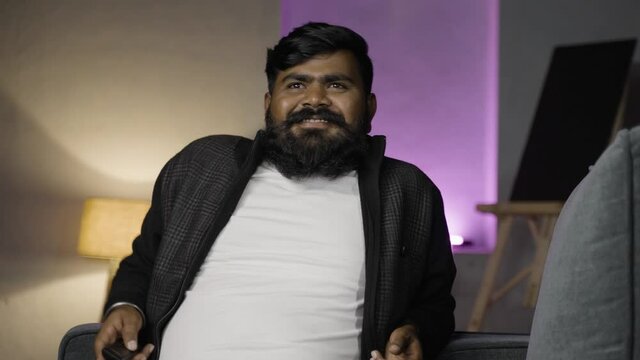 Young indian beard man watching tv or television by changing channal at home - concept of weekend or holiday relaxtion,daily lifestyle and leisure activitys.