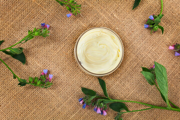 The Common Comfrey (Symphytum officinale) herb with jar of cosmetic cream on a jute background. Top view.