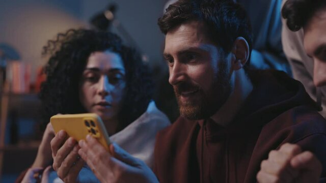 Close up of joyed multiethnic people screaming while watching results of the game on smartphone and sitting on sofa. Focus on bearded guy rejoicing together with friends and screaming.