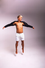 Attractive handsome young blond man wearing black shirt and white shorts jumping for joy in photo studio on light-grey background