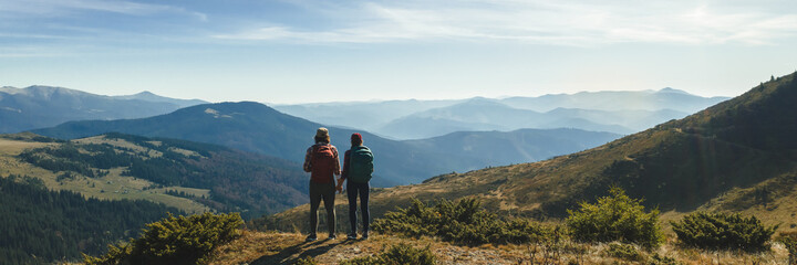 Couple of hikers with backpacks enjoying valley landscape view from top of a mountain. Young adult tourists, man and woman standing on the pass and holding by hands. Panoramic view of mountain hills - Powered by Adobe