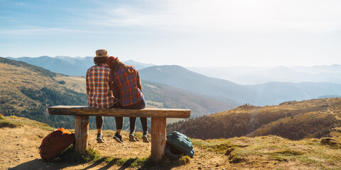 Couple of hikers with backpacks enjoying valley landscape view from top of a mountain. Young adult tourists, man and woman sitting in the bench. Panoramic view of mountain hills, Carpathian mountains - Powered by Adobe