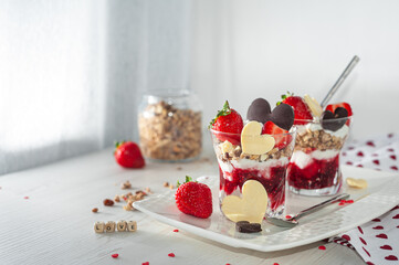 Sweet breakfast on Valentine's Day on a white wooden table. Creamy dessert with ripe strawberries and pureed raspberries, muesli in a glass decorated with chocolate in the shape of a heart. Side view