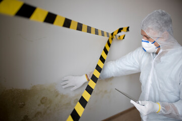 Man with white protective suit and mouth nose mask stands in front of mold on wall and works with tablet behind barrier - obrazy, fototapety, plakaty