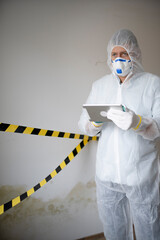 Man with white protective suit and mouth nose mask stands in front of mold on wall and works with...