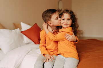 Smiling cute children a little girl and a boy in bright orange pajamas hug and lie on bed linen on a bed in a cozy bedroom at home on a weekend indoor. Selective focus