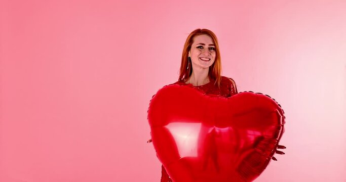 Beautiful redhead girl with red heart balloon posing. Happy Valentine's Day concept. Studio photo of beautiful ginger girl dancing on pink background.