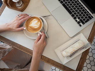 Work life balance. A woman works on a laptop in a cozy cafe with a cup of coffee and an eclair in the morning sun.