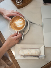 Work life balance. A woman works on a laptop in a cozy cafe with a cup of coffee and an eclair in the morning sun.