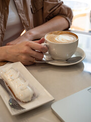 Work life balance. A woman works on a laptop in a cozy cafe with a cup of coffee and an eclair in the morning sun.