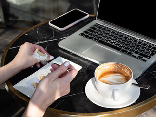 Work life balance. A woman works on a laptop in a cozy cafe with a cup of coffee and an eclair in the morning sun.