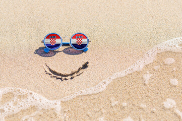Fototapeta na wymiar Sunglasses with flag of Croatia on a sandy beach. Nearby is a sea lightning and a painted smile. The concept of a successful vacation in the resorts of Croatia.