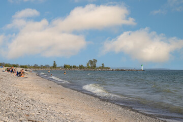 View looking down a rocky and sandy beach along a lakeshore, sunbathers and lighthouse in distance, daytime