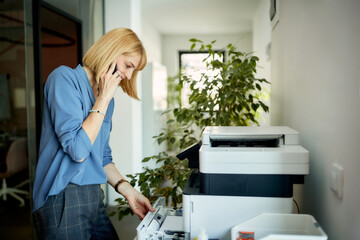 Smiling businesswoman uses photocopier while talking mobile phone in the office.