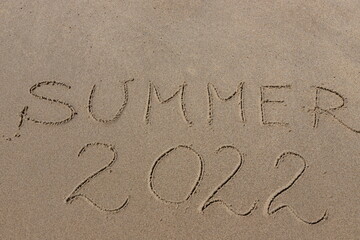The inscription summer 2022 on the sand by the water and the rising wave, seashore beach vacation by the sea