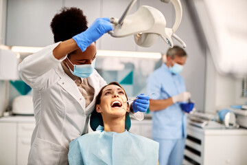 Black female dentist examining teeth of young woman at dental clinic. - obrazy, fototapety, plakaty