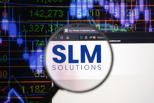 SLM Solutions company logo on a website with blurry stock market developments in the background, seen on a computer screen through a magnifying glass.