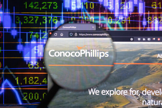 ConocoPhillips company logo on a website with blurry stock market developments in the background, seen on a computer screen through a magnifying glass.