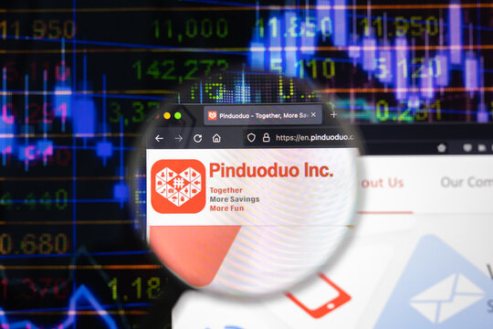 Pinduoduo Inc. company logo on a website with blurry stock market developments in the background, seen on a computer screen through a magnifying glass.