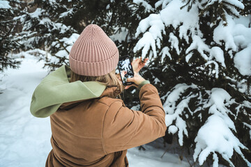 Girl makes a photo on the phone a fir tree with snow in the forest in winter
