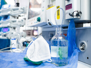 Fototapeta premium A glass bottle with saline and a white medical mask on the table in the hospital against the background of modern medical equipment. Selective focus.