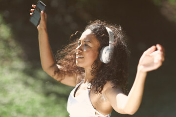Excited girl dancing and listening music with headphones and smart phone in the park with hair moving