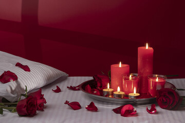 A fragment of a bed in a hotel or at home with a tray with candles, a decoration of red roses and petals. White linens, a red wall with silhouette or shadow. Honeymoon, or Valentine's Day.
