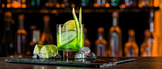 Cocktail with cucumber, ice on bar counter in a restaurant, pub. Fresh tonic drink with lime juice, mint, gin, cucumber juice. Alcoholic cooler beverage at nightclub on dark background