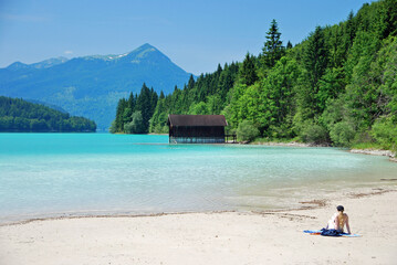 woman sitting on a beach overlooking the turquoise water of  Lake Walchensee in the bavarian alps