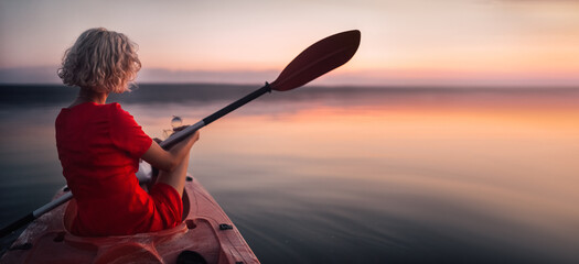 Blonde woman in a red dress sits in a boat with a paddle at sunset.