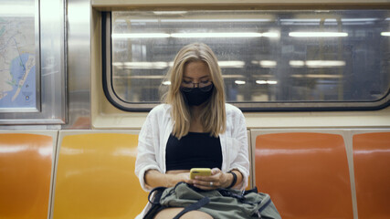 Woman in a black medical face mask to avoid the spread of coronavirus who is sitting alone in a modern subway car. Girl with cellphone in a surgical mask is keeping social distance on a metro train.