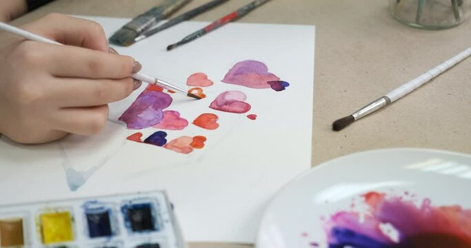 Process of painting card with hearts for Valentine's Day, close-up. Unrecognizable girl drawing a picture, crafting homemade postcard. Desktop with watercolor paint and brushes
