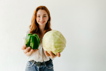 Fototapeta premium Smiling redhead girl with healthy food vegetables, cabbage and green pepper, offering it to the camera. Woman selling organic vegetables at the market