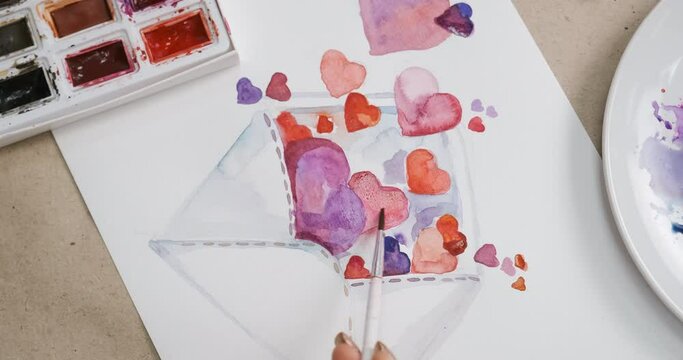 Process of painting card with hearts for Valentine's Day, close-up. Unrecognizable girl drawing a picture, crafting homemade postcard. Desktop with watercolor paint and palette