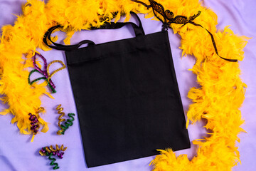 Mardi Gras Mockup black tote bag with glitter decor, carnival mask and yellow feather boa on fabric lilac background, flat lay, top view, copy space.