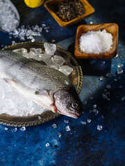 Fresh fish on a plate with ice cubes, ice is scattered on the table, a bowl of salt. Dark blue background. Salting and marinating fish, cooking, fish market, advertising, banner. Close-up.