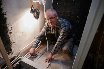 A 59-year-old man in a plaid shirt, wearing glasses figuratively, masterfully lays tiles around the water drain hole in a cheap cabin