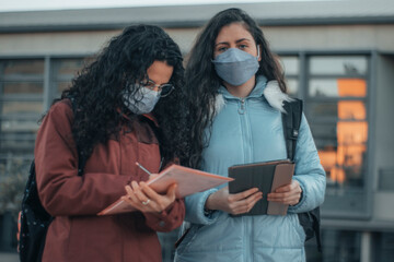 Chicas jóvenes de cabello oscuro concentradas con mascarilla hablando y revisando las tareas y...