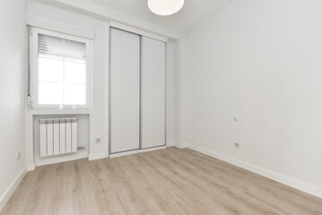 Empty room with light wooden floor, sliding white wooden wardrobe doors and aluminum window above aluminum radiator and ceiling lamp