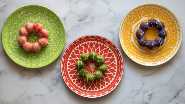 Three colorful plates with a geometric design, with three different Mochi Donuts.  Strawberry, Ube, and Matcha glaze.