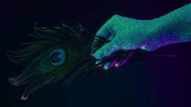 Female hand covered with holographic shining glitter holding peacock feather under neon colored light. Body art, glamorous conceptual footage, fashion style. 