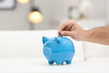 Young man putting money into piggy bank at white table indoors, closeup