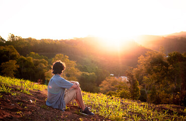 Fototapeta premium Young man sitting on top of a hill and watching the sunset. Person thinking and having reflections