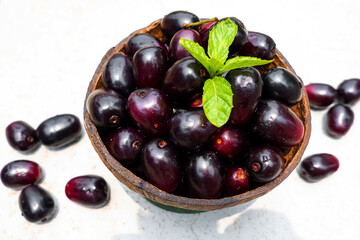 Dark pink-red ripe Syzygium cumini fruits. Dark black java plum in a wood bowl at isolated white background. Green mint leaf on top of some large java plums.