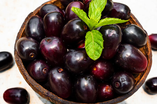 Dark pink-red ripe Syzygium cumini fruits. Dark black java plum in a wood bowl at isolated white background. Green mint leaf on top of some large java plums.