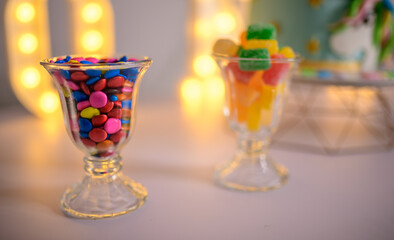 Birthday party sweets and treats, decorated lighting background. a glass ful of multicolored chocolate pebbles and jujubes.