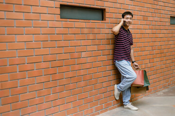 shopping concept The guy in a happy face attempting to make a phone call while slanting himself on the wall and bringing plenty of shopping bags with him